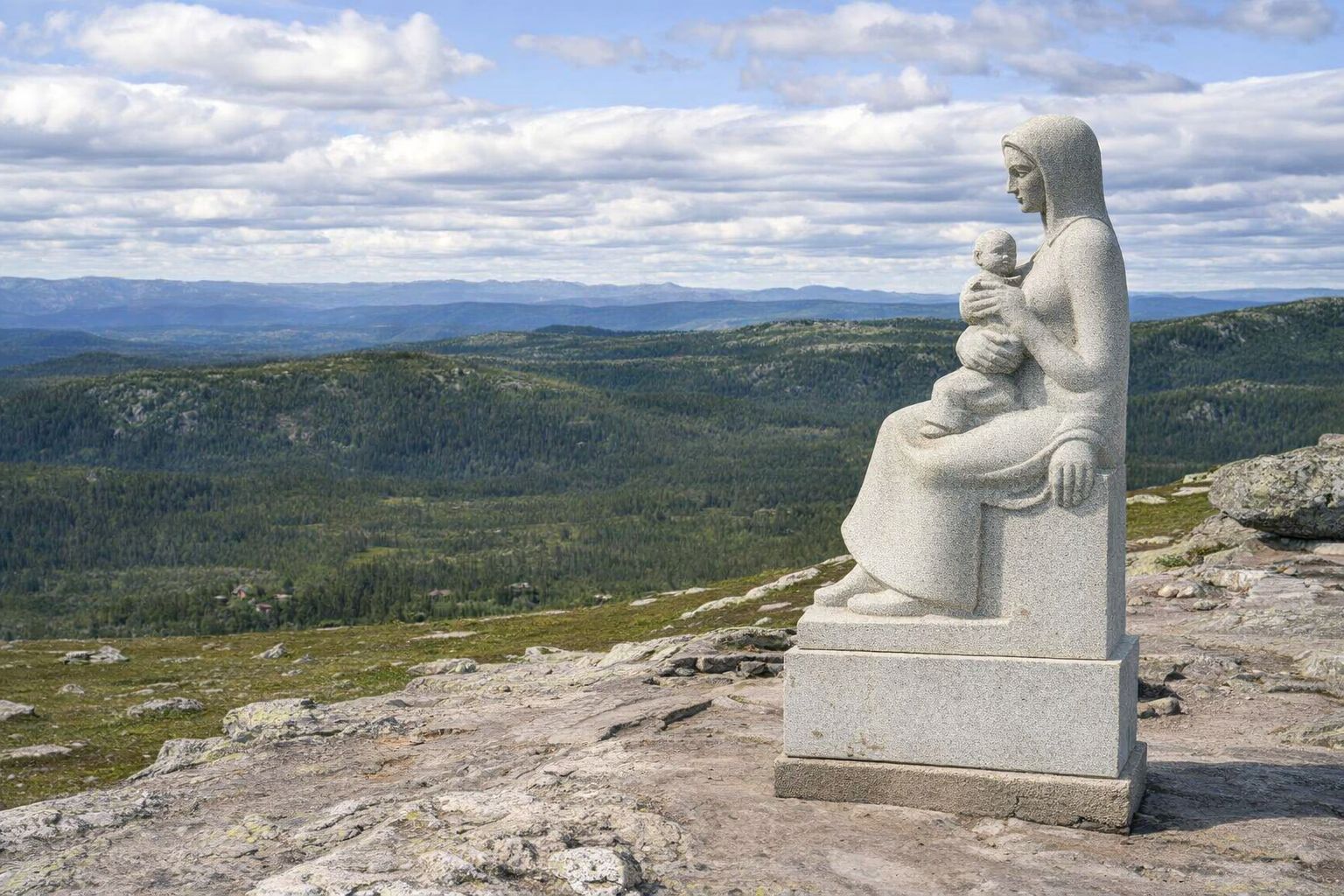 Stenskulptur av en mor och barn på ett berg med utsikt över skogslandskap.